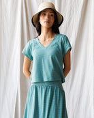 Woman wearing a teal striped tee and skirt and hat against a white curtain background
