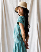 Woman wearing a teal dress and straw hat against a white curtain background