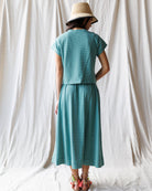 Person wearing a teal dress and hat standing against a white curtain