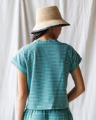 Person wearing a teal striped outfit with a straw hat against a white curtain background