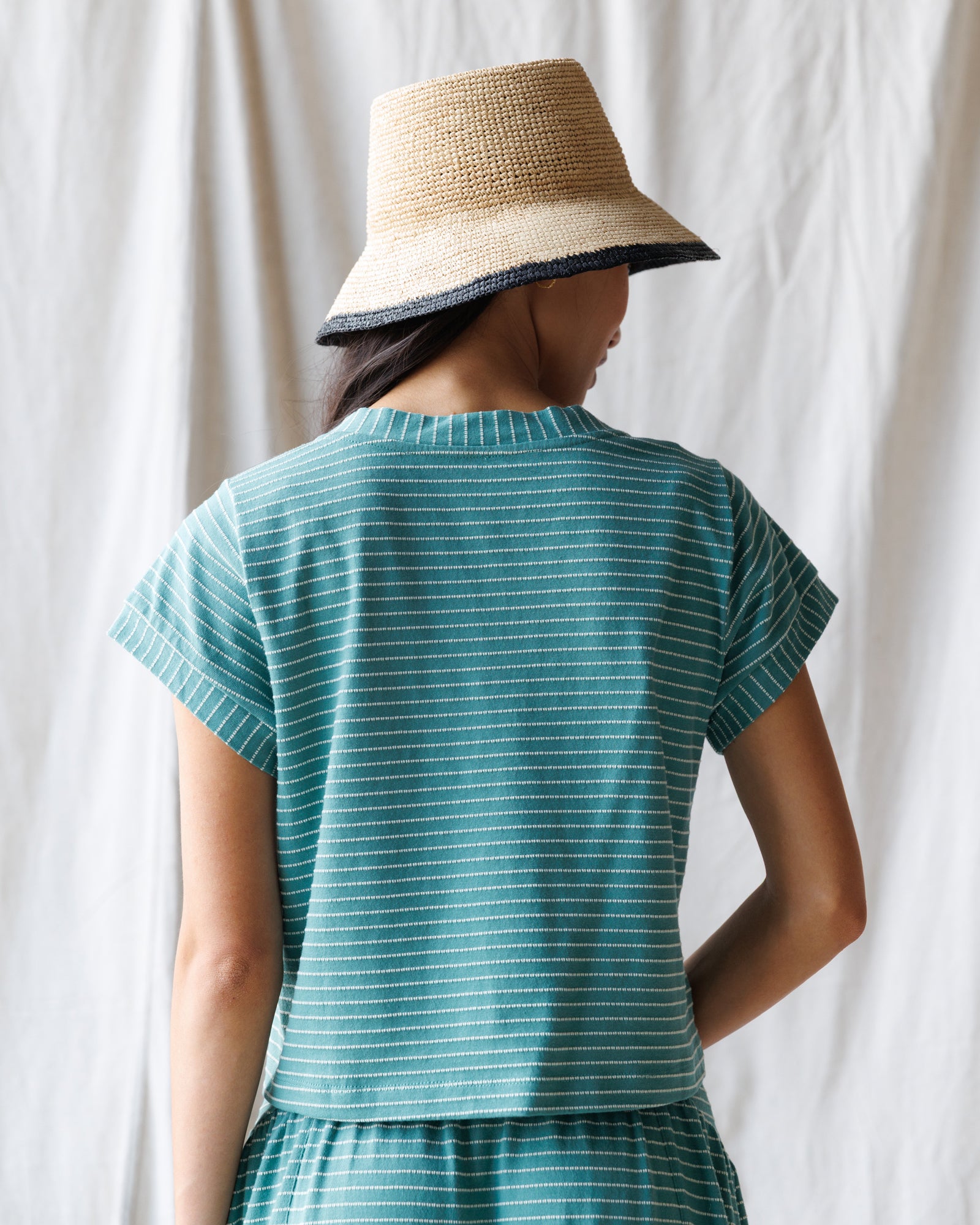 Person wearing a teal striped outfit with a straw hat against a white curtain background