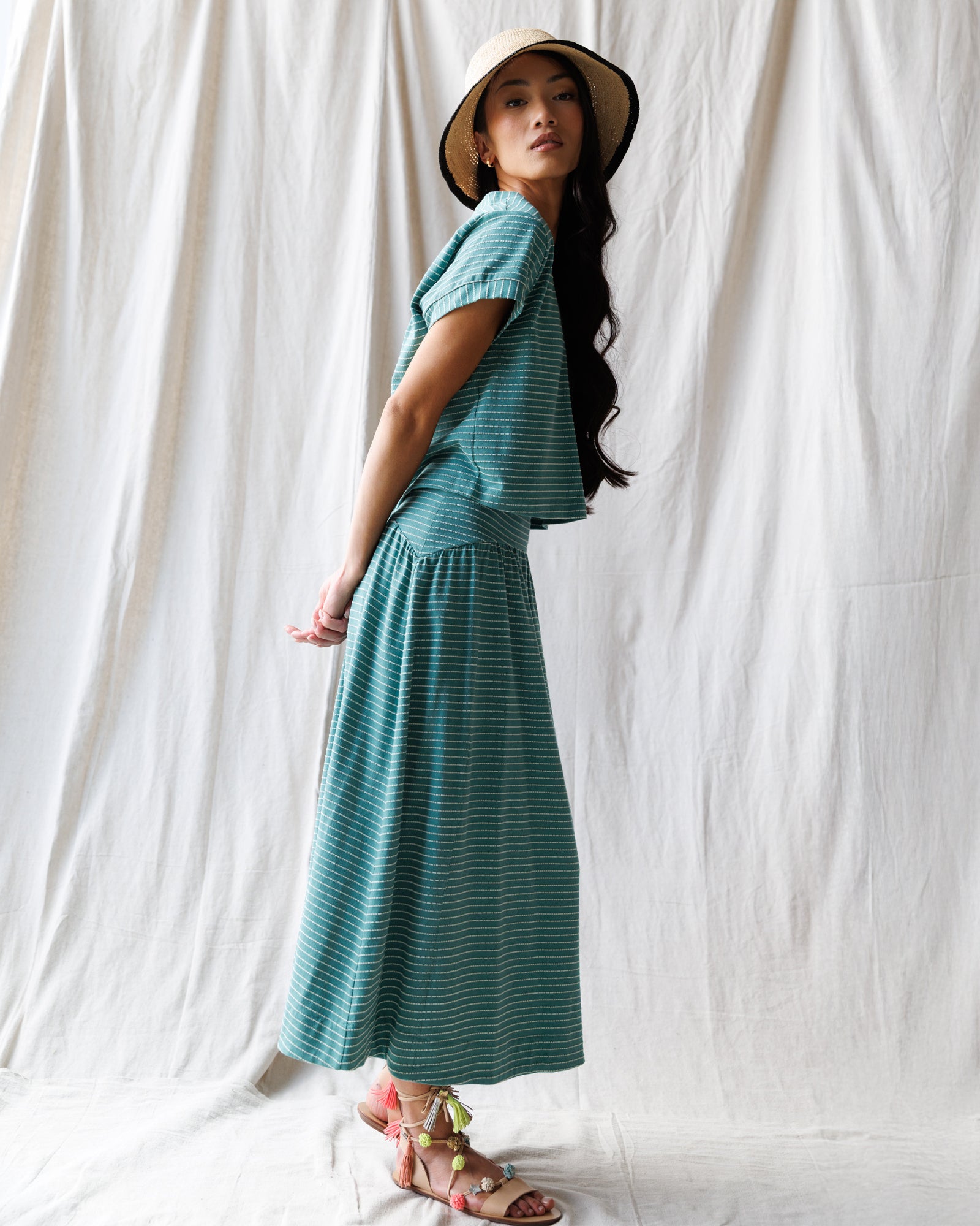 Woman wearing a teal striped dress and hat against a white curtain background