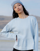 A woman wearing a light blue crewneck sweatshirt with a peplum waist and a matching light blue beanie, photographed outdoors against a soft-focus natural background.
