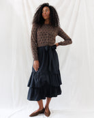 A-line black poplin midi skirt featuring an optional matching self-tie sash at the waist and two full, gathered flounce tiers, styled with a brown polka-dot cardigan.