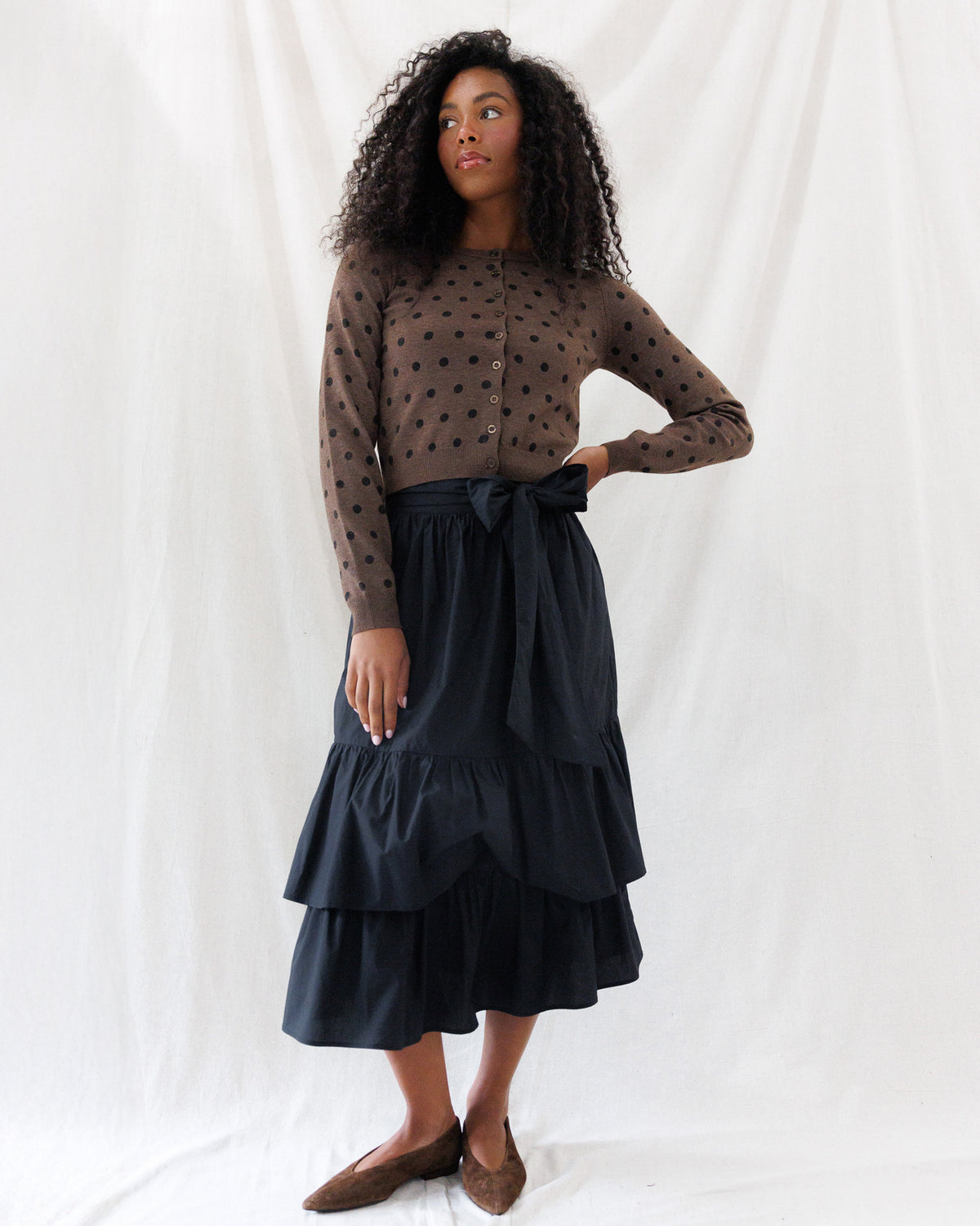A-line black poplin midi skirt featuring an optional matching self-tie sash at the waist and two full, gathered flounce tiers, styled with a brown polka-dot cardigan.