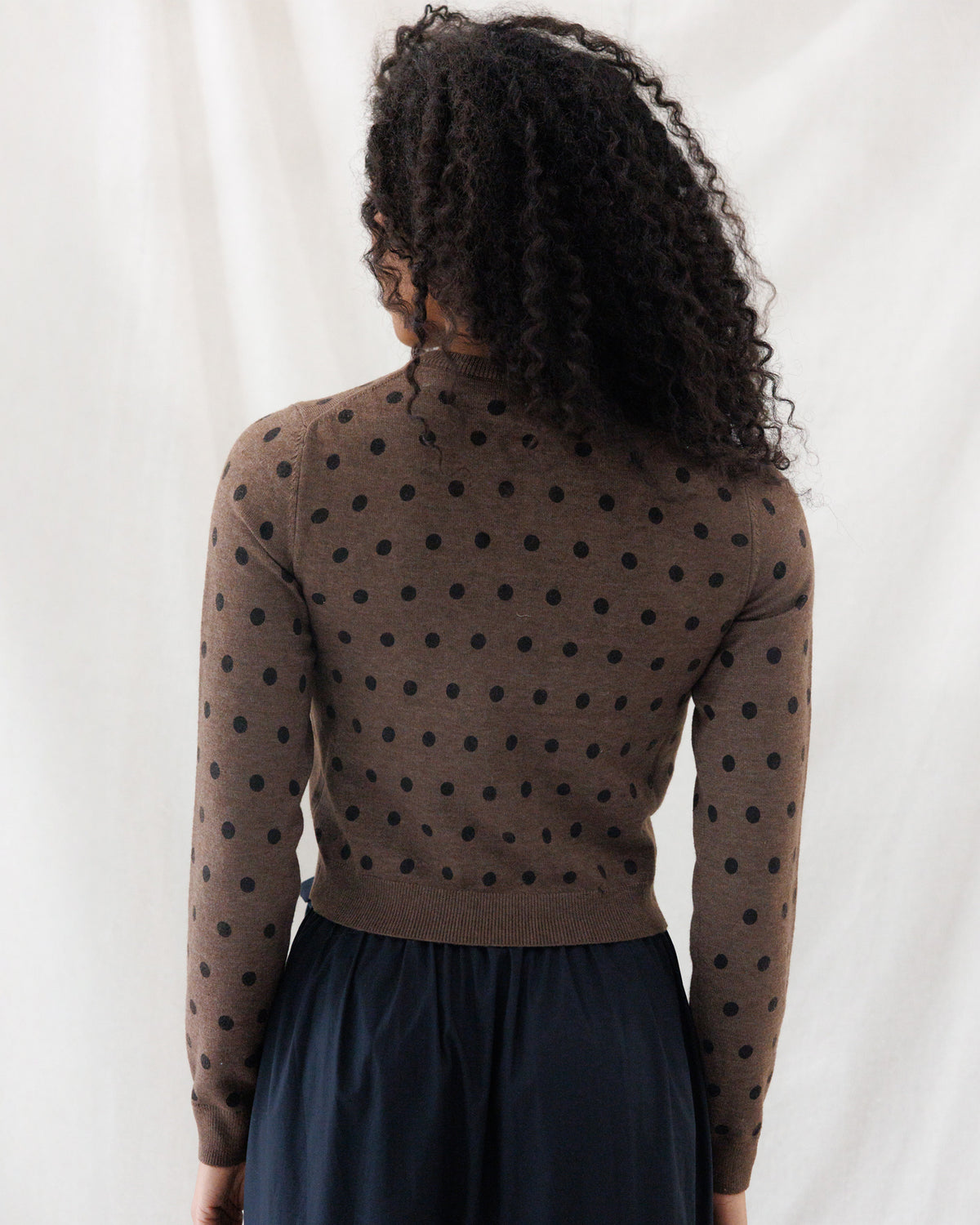 A person shown from the back wearing a brown long-sleeve knit top with black polka dots and a dark navy skirt, photographed against a white draped fabric background.