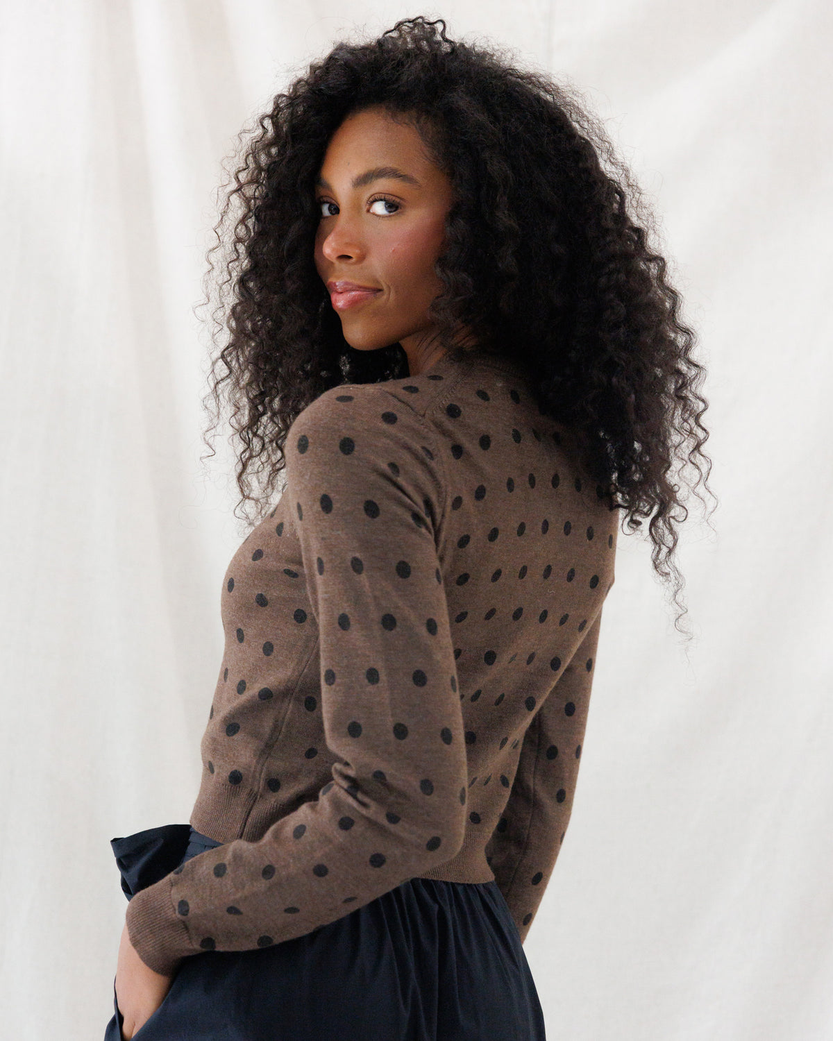 A woman with long, curly black hair is shown from the back wearing a brown long-sleeve knit top with black polka dots, paired with a dark navy skirt, against a white draped fabric background.