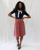 A woman stands against a white draped fabric backdrop wearing a dark navy short-sleeve top with a white lace tie at the collar, a red and green plaid pleated knee-length skirt, and dark lace-up flats.