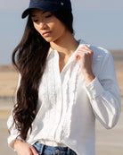 A woman wearing a white long-sleeve button-up shirt with ruffle details, blue jeans, and a navy baseball cap, photographed outdoors against a blurred desert-like background.