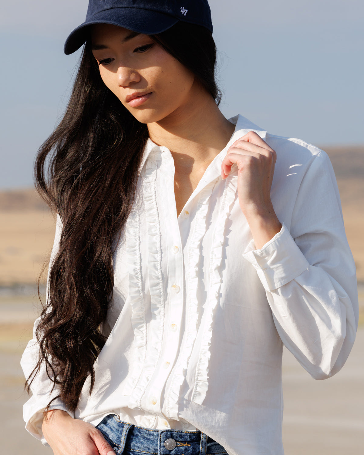 A woman wearing a white long-sleeve button-up shirt with ruffle details, blue jeans, and a navy baseball cap, photographed outdoors against a blurred desert-like background.
