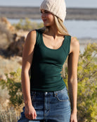 A woman wearing a dark green scoop-neck tank top, blue jeans, and a cream knit beanie, standing outdoors in a natural, rocky environment.