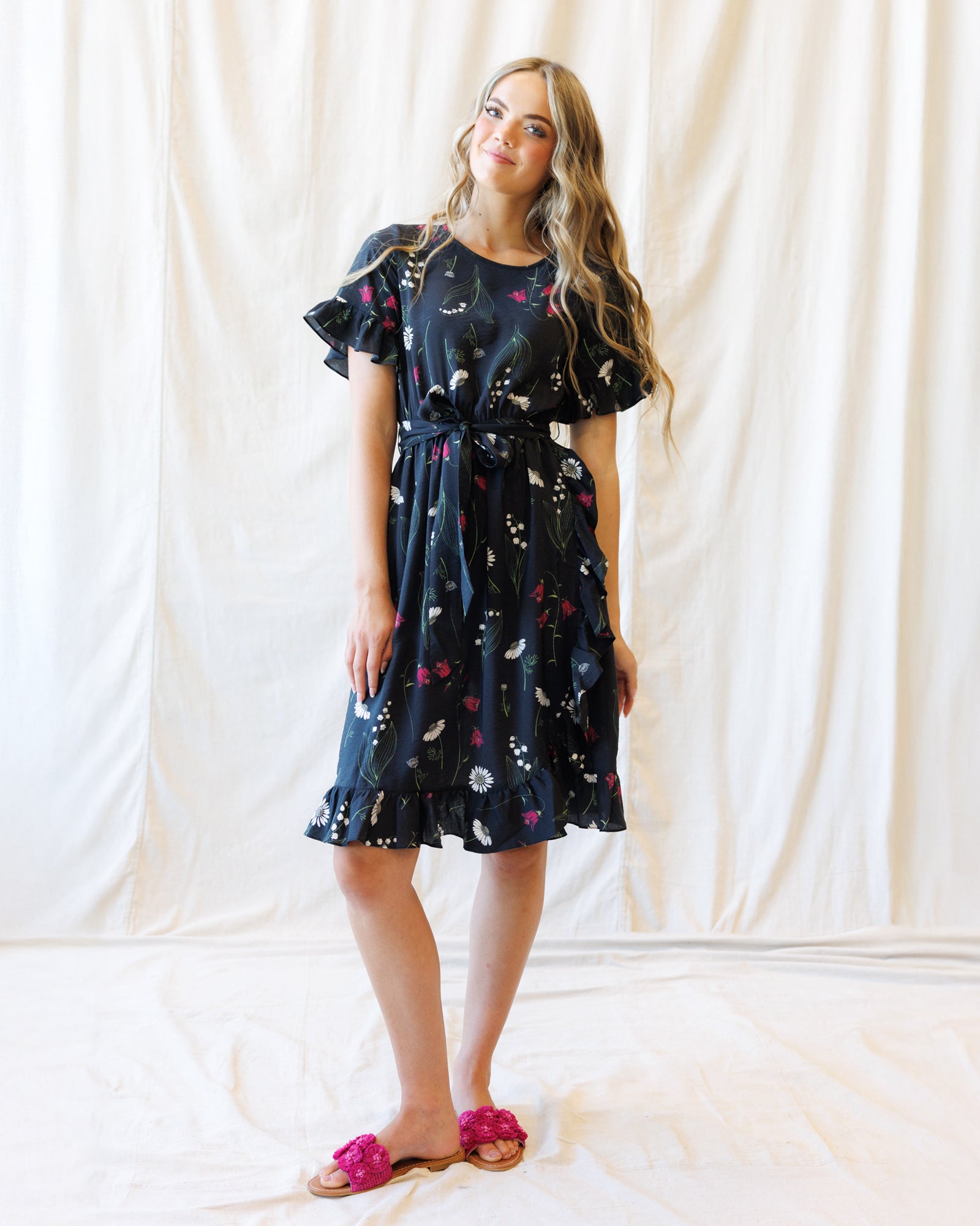 A woman modeling a dark navy blue floral print dress with ruffled short sleeves and a matching tie belt, standing against a draped white fabric backdrop.