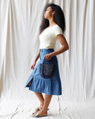 Woman wearing a white top and blue denim skirt holding a blue handbag against a white background