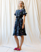 A woman models a dark navy blue short-sleeve dress with a colorful abstract print, featuring a tie waist and ruffled sleeves, paired with pink slide sandals against a draped cream fabric backdrop.