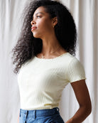 Woman wearing a light-colored top and blue pants against a white curtain background