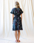 A woman viewed from behind wearing a dark navy blue knee-length dress with a white and pink floral print, standing against a draped cream fabric backdrop.