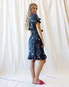 A woman modeling a short-sleeve navy blue dress with a white and pink abstract print, standing against a draped cream fabric backdrop.