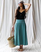 Woman wearing a black top and teal skirt holding a straw bag against a white curtain background