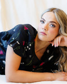 A woman with blonde hair lies on a white surface, wearing a black short-sleeve blouse with a colorful floral and leaf pattern.