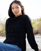 A woman wearing a black long-sleeve top with a smocked bodice and peplum hem, paired with blue jeans, sitting outdoors on a sandy surface with dry vegetation in the background.