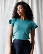 Woman wearing a teal top with ruffled sleeves against a white curtain background