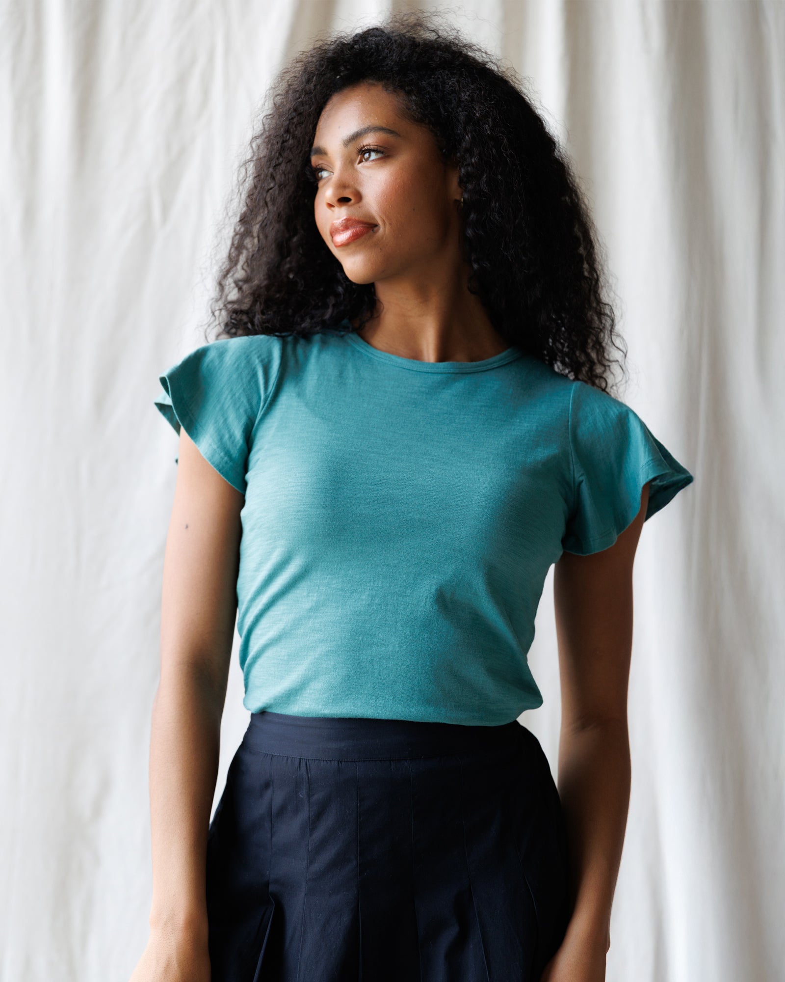 Woman wearing a teal top with ruffled sleeves against a white curtain background