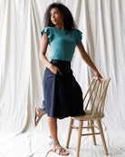 Woman in teal top and black skirt standing next to a wooden chair against a white curtain background