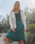 A blonde woman wearing a cream cardigan, dark green tank top, and a dark green pleated skirt, standing outdoors in a sandy, brushy environment.
