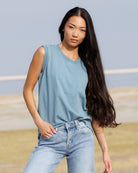 A woman with long dark hair wearing a light blue sleeveless crewneck top and light-wash blue jeans, standing outdoors against a soft-focus background of sky and grass.