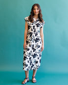 A woman modeling a white midi dress with a black floral print, standing against a solid teal background.