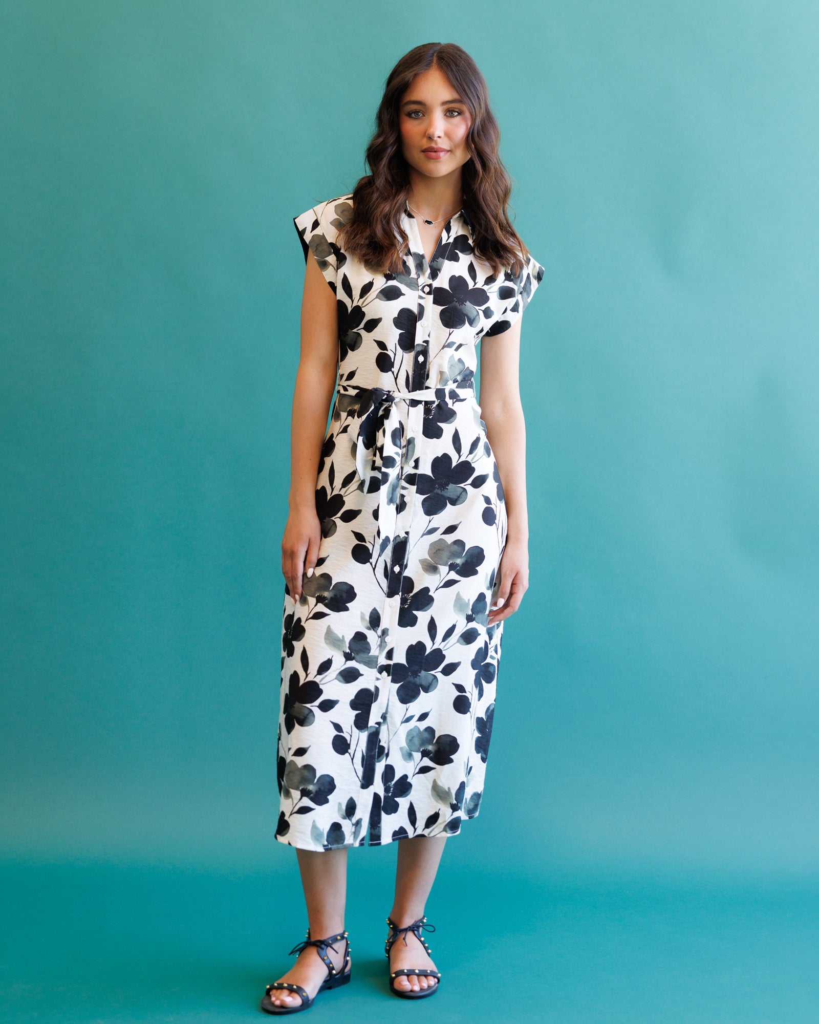 A woman modeling a white midi dress with a black floral print, standing against a solid teal background.