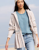 A woman wearing a light beige hooded cardigan with lace trim over a dusty blue t-shirt and light-wash jeans, standing outdoors against a pale sky background.