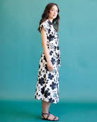 A woman modeling a sleeveless midi dress with a black floral print on a white background, standing against a solid teal studio backdrop.