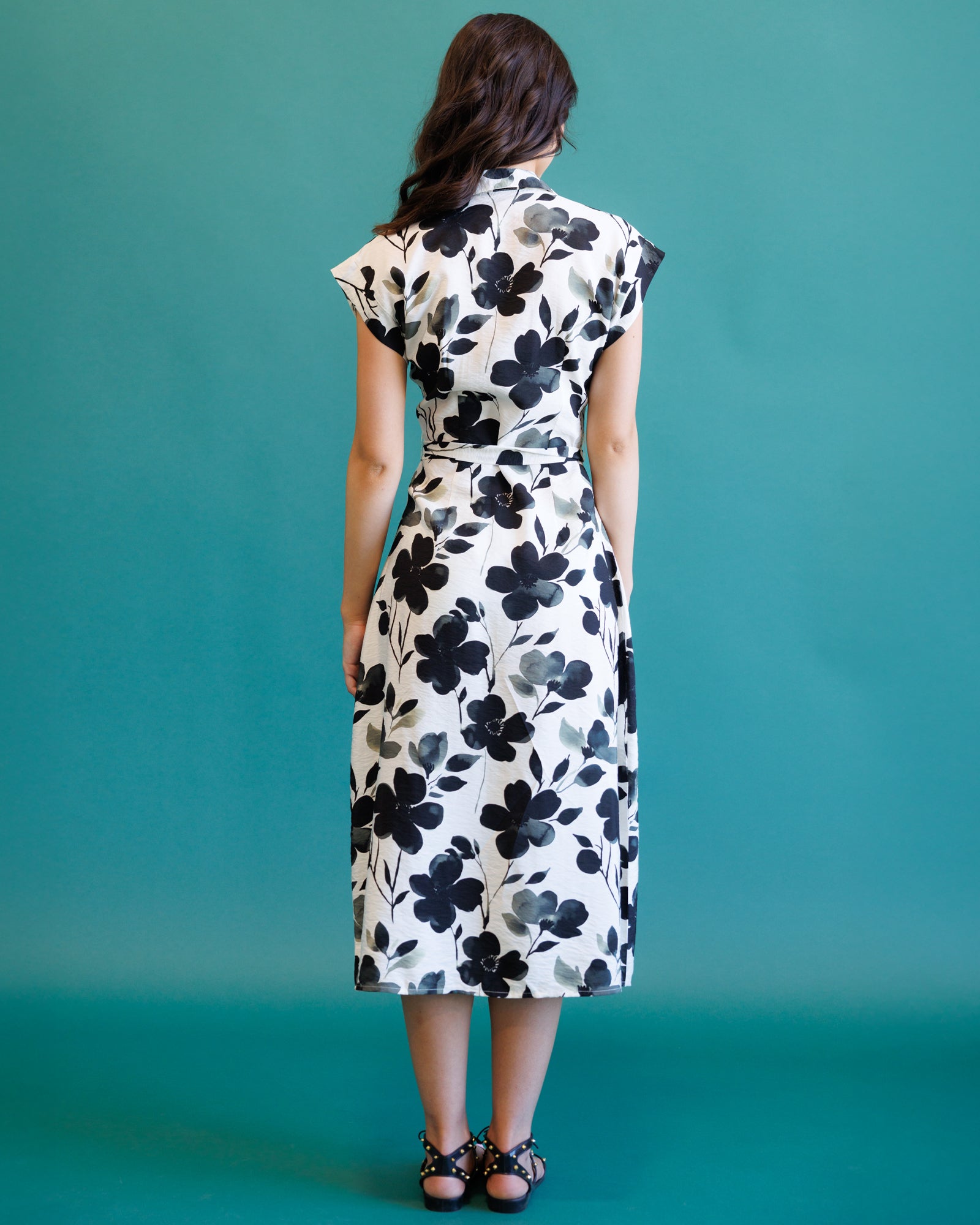 A woman viewed from behind wearing a knee-length white dress with a bold black floral print, a high collar, and cap sleeves, standing against a solid teal background.