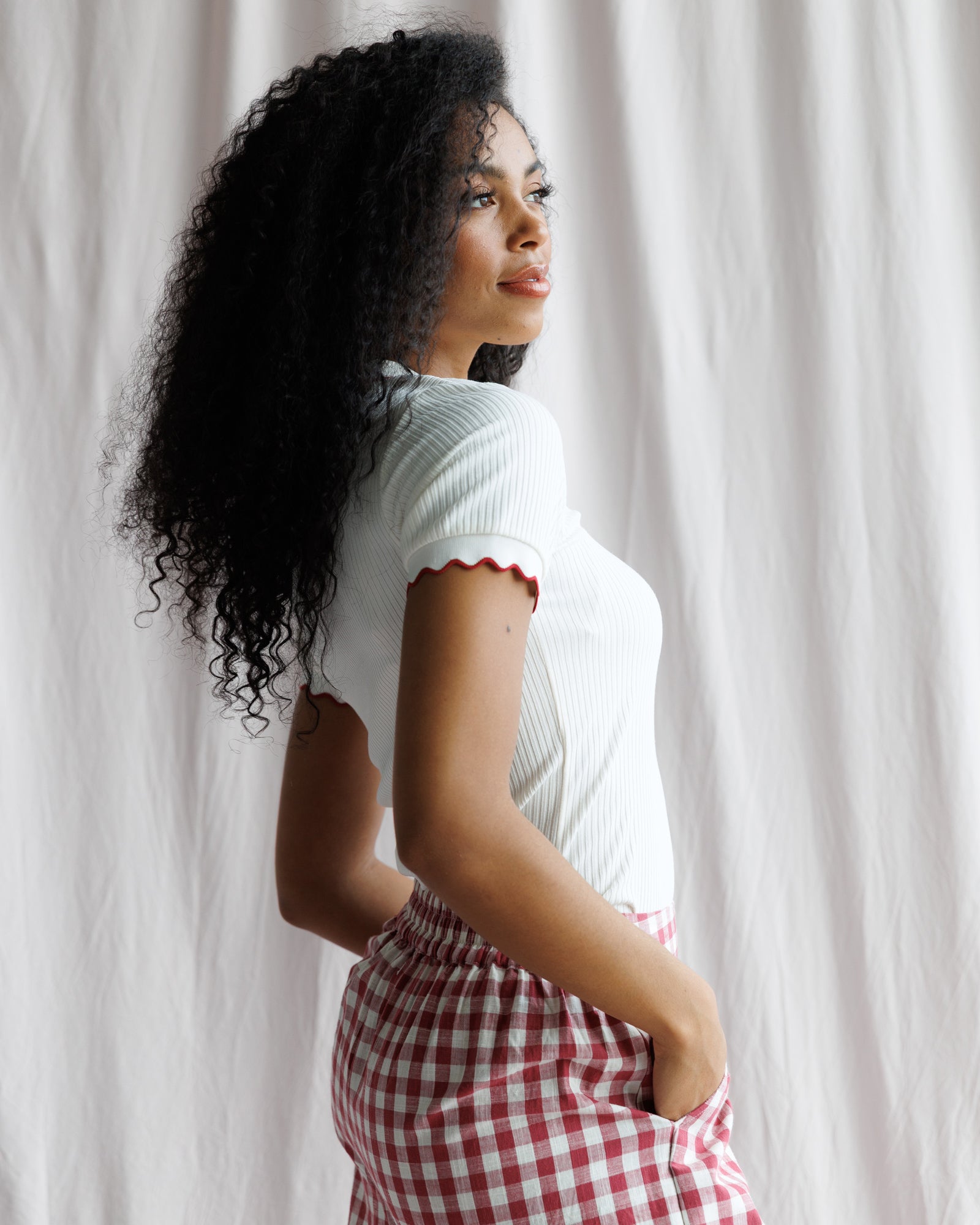Woman standing against a white curtain wearing a white top and red checkered skirt.