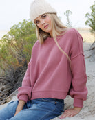 A woman wearing a dusty rose ribbed knit crewneck sweater, blue jeans, and a cream cable-knit beanie sits on a sandy beach with shrubs in the background.