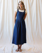 A woman stands against a draped cream fabric backdrop wearing a dark blue denim pinafore-style dress over a short-sleeve cream top, with metallic sandals.