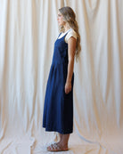 A woman stands in profile wearing a dark blue denim pinafore dress over a white short-sleeve top, standing against a draped cream fabric backdrop.