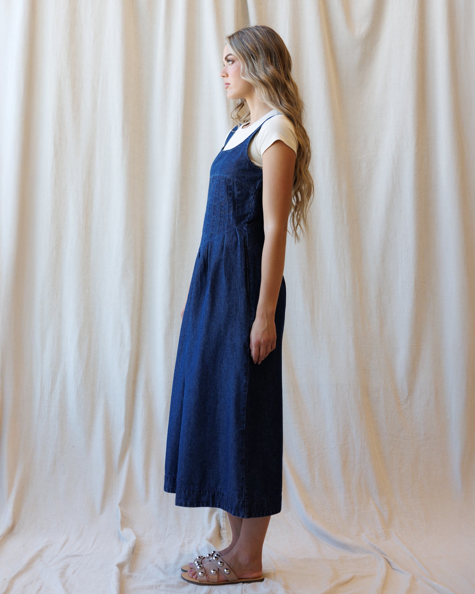 A woman stands in profile wearing a dark blue denim pinafore dress over a white short-sleeve top, standing against a draped cream fabric backdrop.