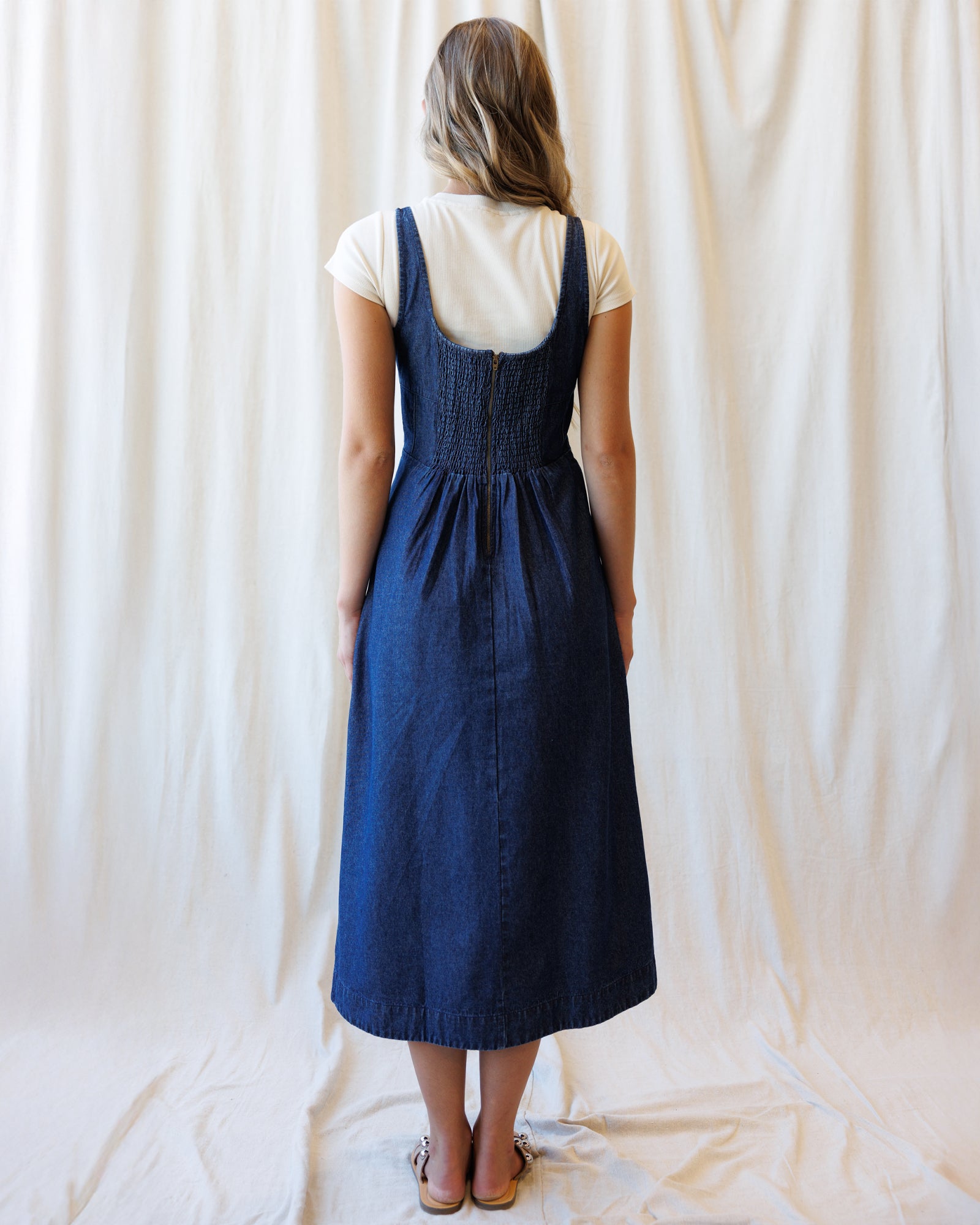 A woman stands with her back to the camera wearing a dark blue denim pinafore dress over a cream short-sleeve t-shirt, set against a draped cream fabric backdrop.
