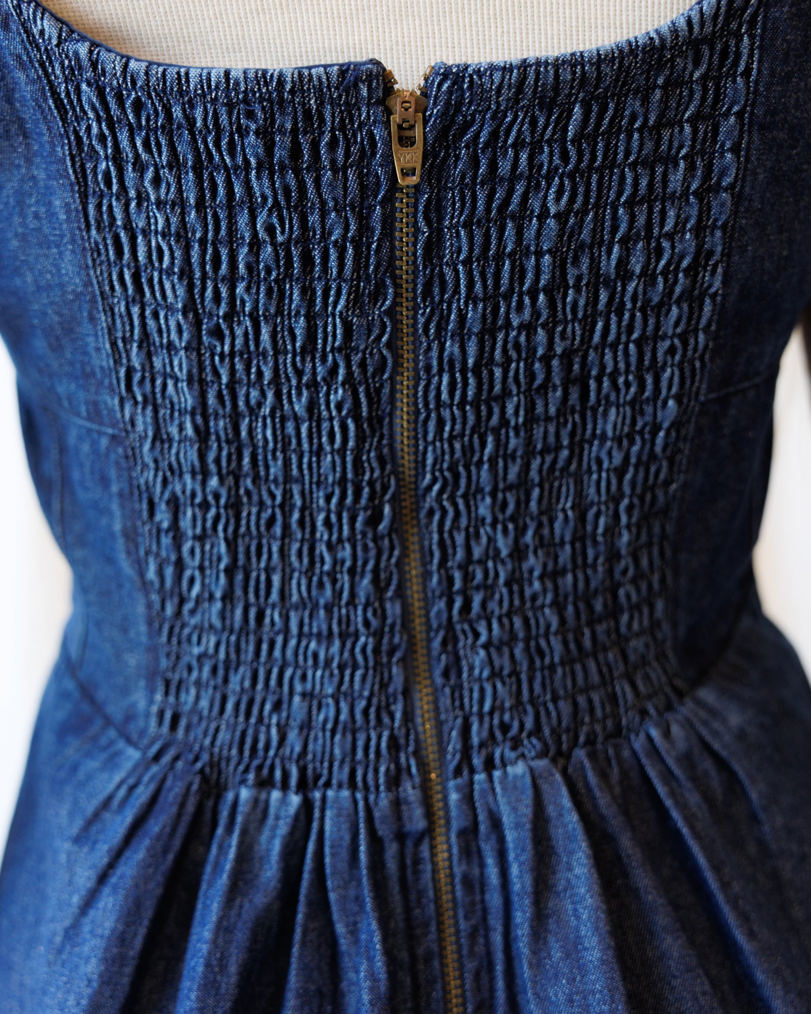 Close-up of a dark blue denim dress with a smocked bodice and a central gold zipper, shown on a mannequin torso against a light background.