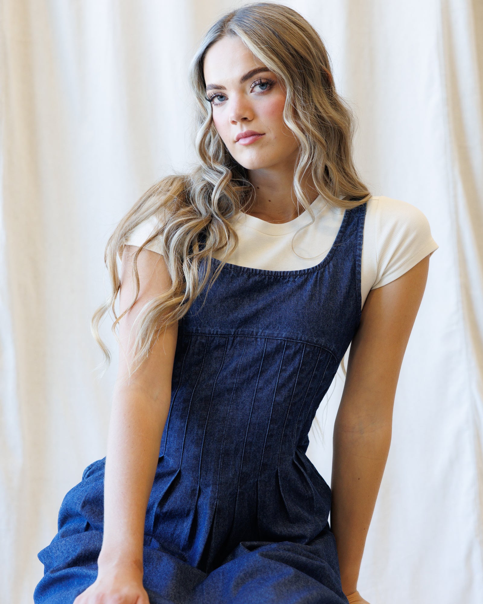 A woman with long wavy blonde hair wearing a dark blue denim overall jumpsuit over a cream short-sleeve t-shirt, posed against a soft, draped off-white fabric background.