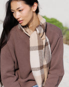 A woman wearing a brown waffle-knit sweater and a beige and brown plaid scarf, photographed outdoors with a soft-focus light background.