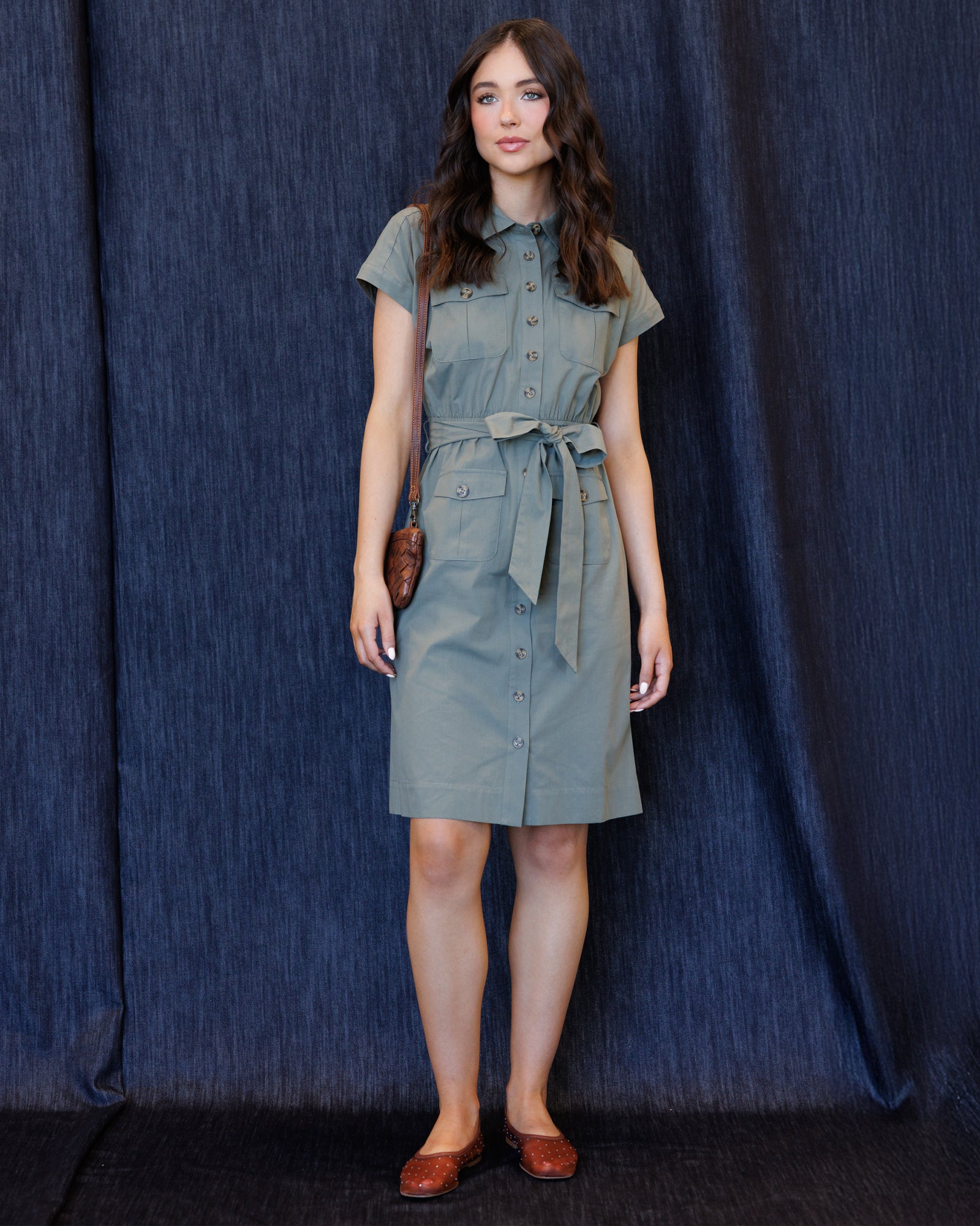 A woman stands against a dark blue textured backdrop wearing a short-sleeve button-front dress with a tie waist and brown shoes.