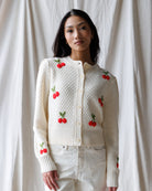 Woman wearing a white knitted cardigan with red cherry designs against a plain background