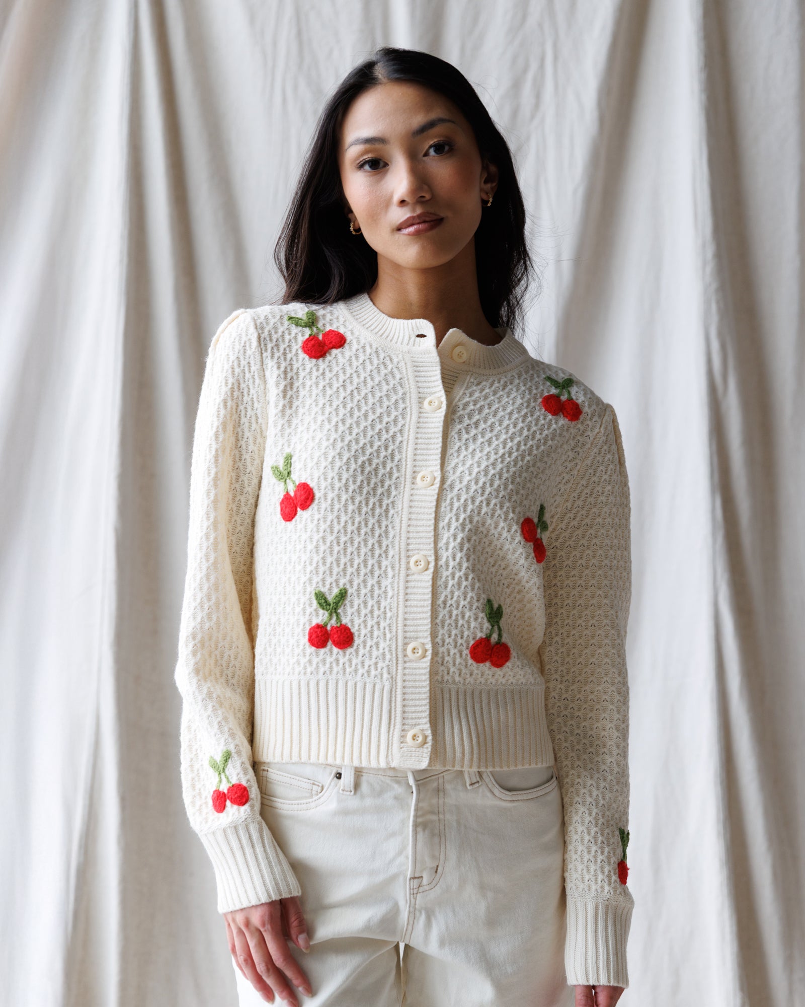 Woman wearing a white knitted cardigan with red cherry designs against a plain background