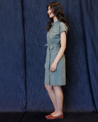 A woman stands in profile wearing a short-sleeve collared dress with a tie waist, photographed against a dark blue textured curtain backdrop.