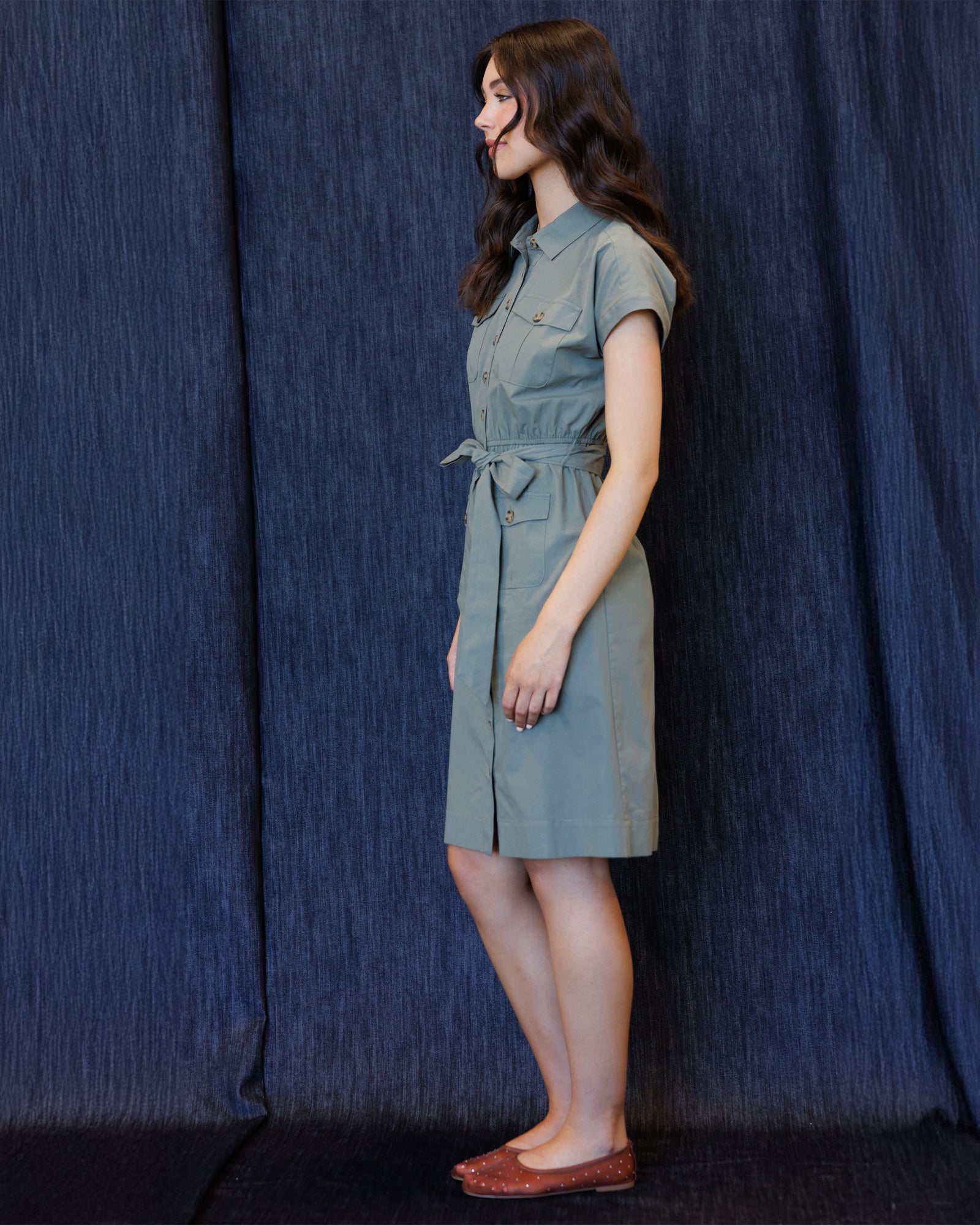 A woman stands in profile wearing a short-sleeve collared dress with a tie waist, photographed against a dark blue textured curtain backdrop.