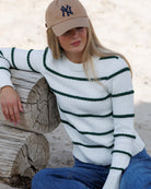 A woman wearing a white knit sweater with dark green horizontal stripes, blue jeans, and a tan baseball cap, sitting in a sandy area with their arm on a log