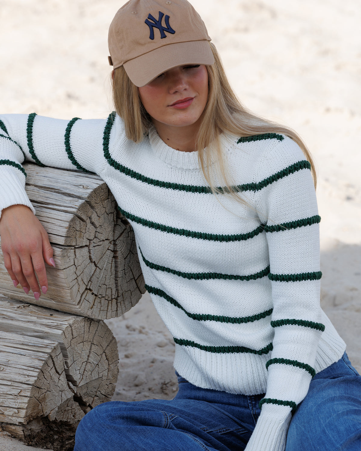 A woman wearing a white knit sweater with dark green horizontal stripes, blue jeans, and a tan baseball cap, sitting in a sandy area with their arm on a log